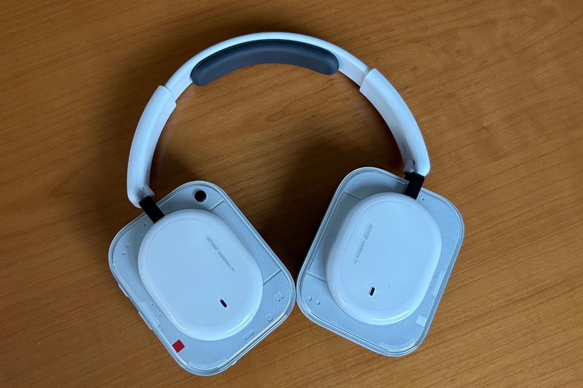 Nothing Headphone (a) White aperte e viste dall'alto: i due padiglioni mostrano il design semi-trasparente con cap ovale centrale, archetto con imbottitura grigia e lo snodo laterale in metallo nero. (mistergadget.tech)