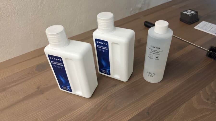 Tre flaconi di detergente Dreame su piano in legno: due bianchi da 500ml Multi-Surface Floor Cleaner e uno trasparente da 250ml Spot Formula for Carpet, con spazzolino di pulizia sullo sfondo