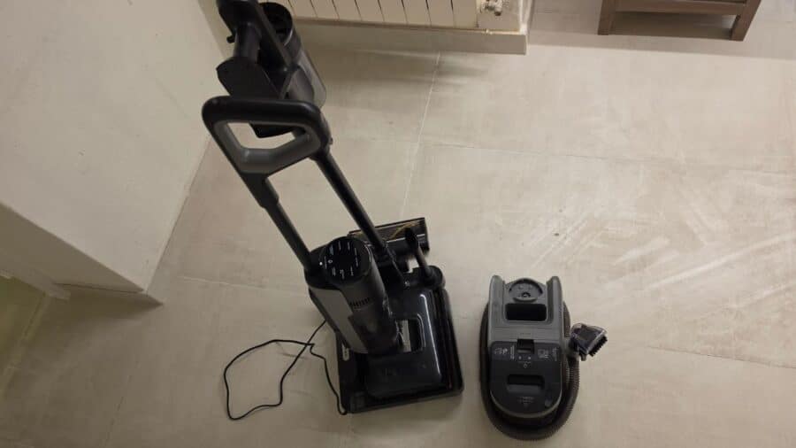 Vista dall'alto del sistema Dreame H15 Mix sul pavimento: a sinistra la lavapavimenti verticale agganciata alla stazione di ricarica nera con cavo, a destra il modulo lavatappeti grigio con tubo flessibile e accessori