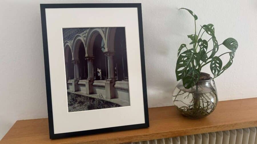 Una cornice SwitchBot AI Art Frame appoggiata su una mensola in legno mostra la foto di un porticato ad archi; accanto c’è un vaso di vetro con una pianta di monstera.