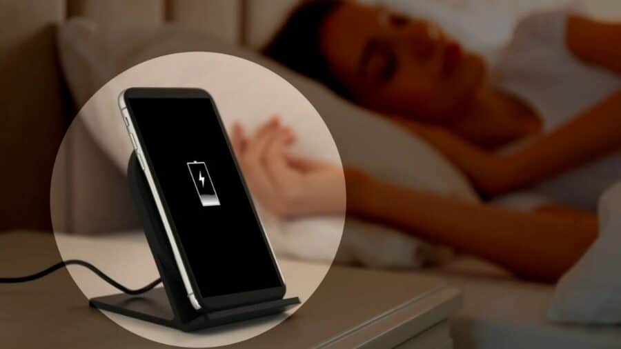 Uno smartphone in carica su una base wireless è appoggiato sul comodino mentre una persona dorme sullo sfondo.