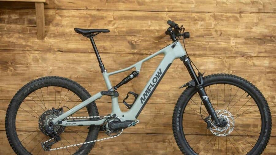 Una e-bike Amflow grigia è appoggiata davanti a una parete in legno, mostrando il telaio full suspended e la configurazione da trail elettrico.