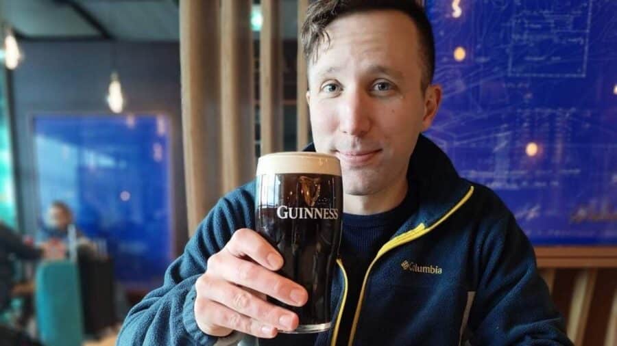 Un uomo seduto in un pub solleva un bicchiere di Guinness pieno con schiuma compatta guardando verso la fotocamera.