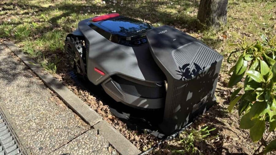 Vista laterale del MOVA LiDAX Ultra 800 parcheggiato sulla base di ricarica tra prato, foglie secche e piante da giardino.