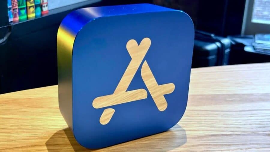 Grande icona tridimensionale dell’App Store Apple appoggiata su un tavolo in legno, con finitura blu e simbolo stilizzato chiaro in primo piano.