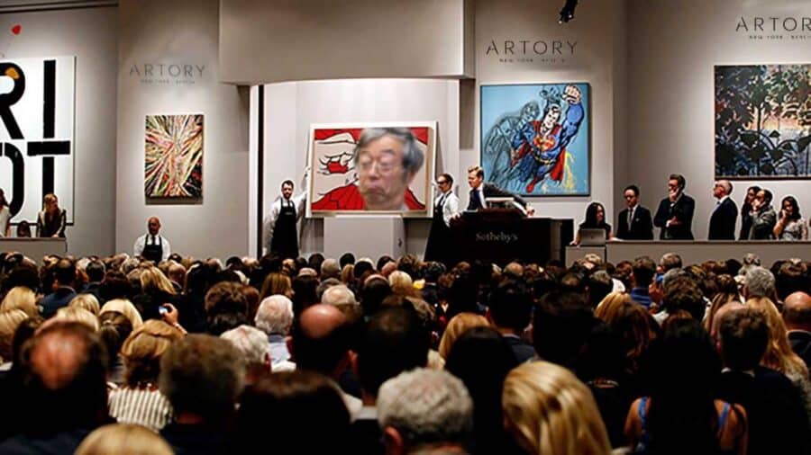 Una sala d’aste gremita osserva un banditore sul palco davanti a grandi opere d’arte esposte alle pareti.