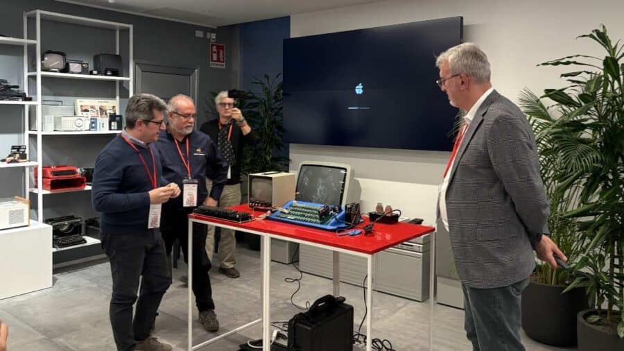 Apple I seriale 002 su tavolo rosso con monitor vintage, tre persone osservano durante l'evento Magneti Media Network