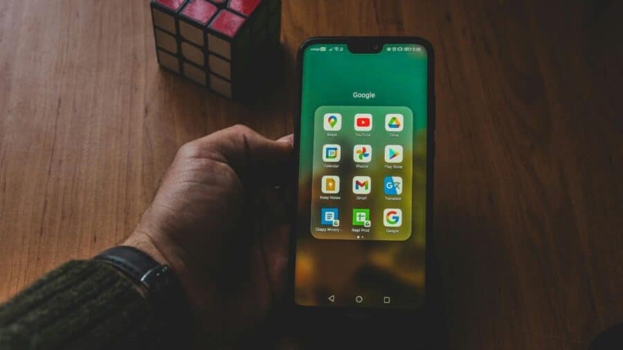 Mano che regge uno smartphone Android con cartella Google aperta, su un tavolo di legno con un cubo di Rubik sullo sfondo