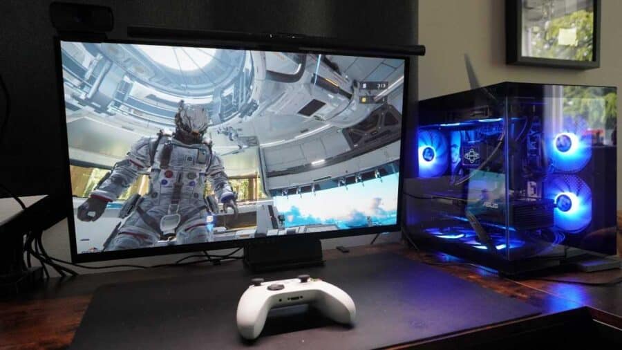 Monitor da gaming che mostra una scena di Pragmata accanto a un PC desktop con ventole RGB blu e controller bianco appoggiato sulla scrivania.