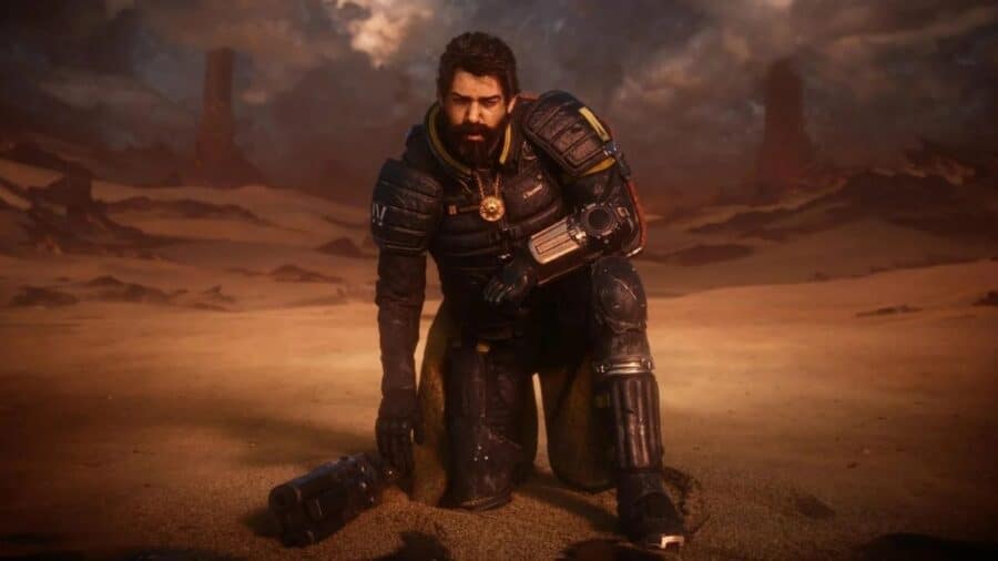 Il protagonista di Saros inginocchiato sulla sabbia di un paesaggio desertico alieno, con armatura futuristica scura e arma appoggiata a terra.