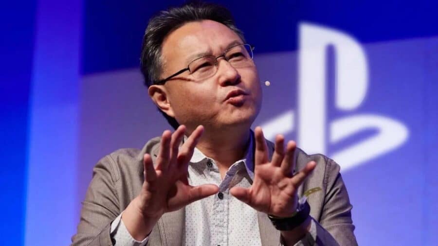 Shuhei Yoshida durante un intervento pubblico su un palco PlayStation, in primo piano mentre gesticola con le mani davanti al logo Sony sullo sfondo blu.