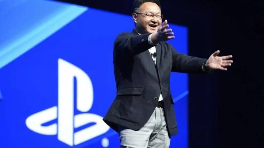 Shuhei Yoshida sorridente sul palco di un evento PlayStation, con le braccia aperte davanti a un grande logo del brand proiettato sullo sfondo.