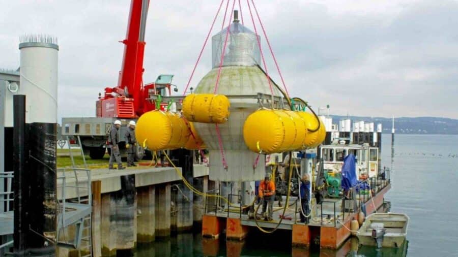 Una grande sfera di cemento con galleggianti gialli viene sollevata da una gru e calata in mare durante una fase di installazione vicino a un molo.