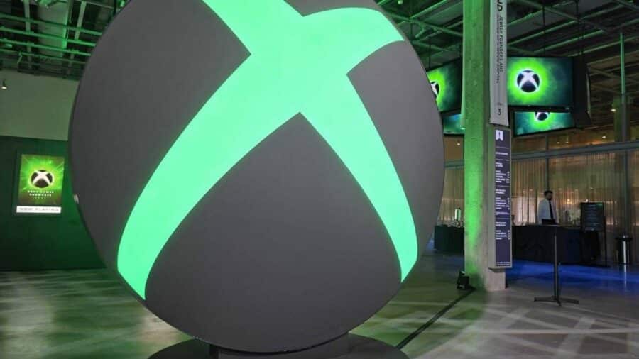Grande installazione luminosa con il logo Xbox verde all’interno di uno spazio espositivo, circondata da schermi e pannelli dedicati al brand Microsoft Gaming.