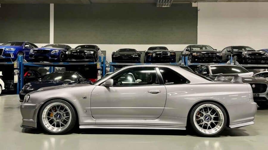 Nissan Skyline GT-R R34 argento vista di profilo all’interno di uno showroom, con assetto ribassato e cerchi sportivi multirazze.