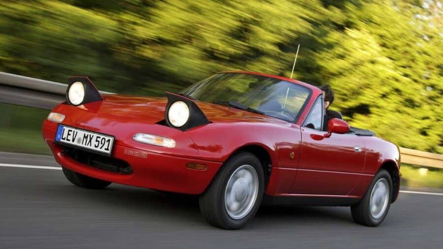 Mazda MX-5 Miata NA rossa con fari a scomparsa aperti, fotografata in movimento su strada con il tetto abbassato.