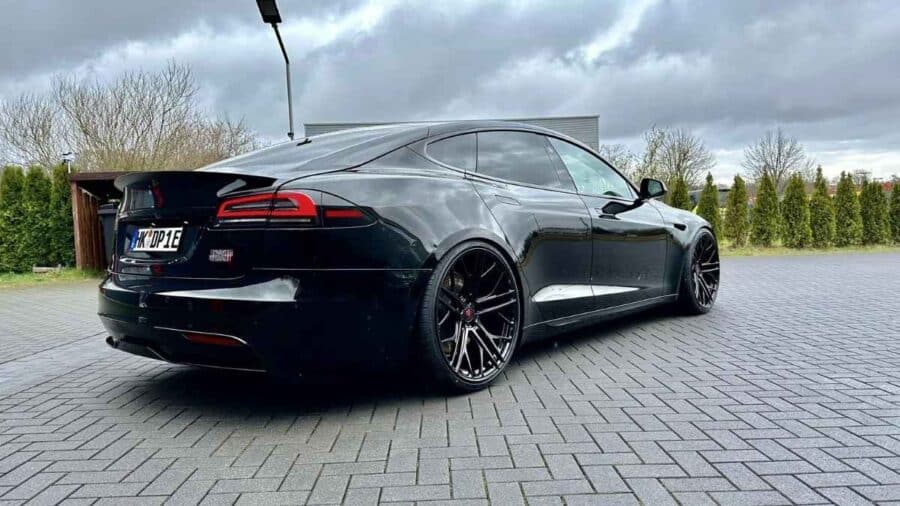 Tesla Model S Plaid nera vista da dietro e di lato, parcheggiata su pavimentazione bagnata con grandi cerchi aftermarket neri.