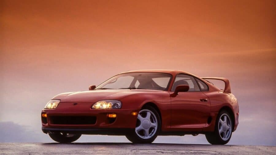 Toyota Supra Mk4 rossa fotografata di tre quarti anteriore su sfondo neutro al tramonto, con alettone posteriore integrato e linea da coupé anni ’90.