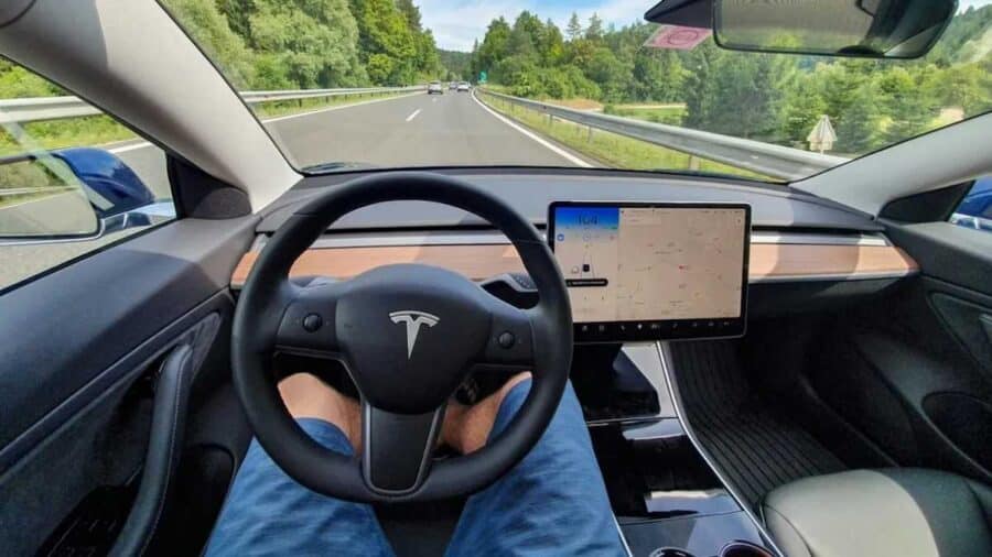 Vista dall’abitacolo di una Tesla in marcia su una strada extraurbana, con volante, display centrale acceso e carreggiata libera davanti al veicolo.