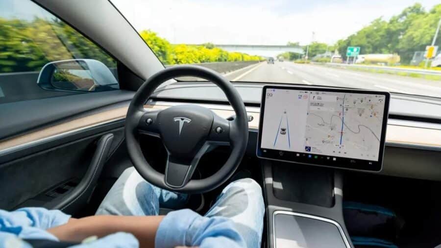 Interno di una Tesla su autostrada, con il grande schermo centrale che mostra navigazione e corsia, mentre l’auto procede su un tratto veloce a più corsie.