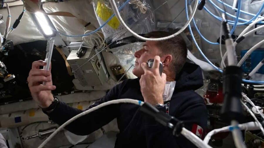 Un astronauta all’interno della capsula spaziale usa uno smartphone circondato da cavi e strumenti di bordo in microgravità.