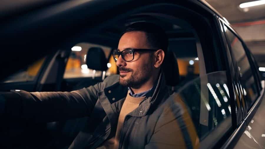 Uomo con occhiali alla guida di un’auto in un parcheggio o ambiente urbano notturno, illuminato dalle luci artificiali dell’abitacolo e della strada.