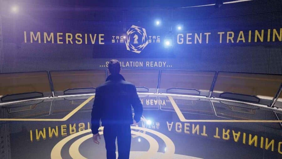 James Bond entra in una sala di addestramento immersiva con grande schermo luminoso e simulazione tattica pronta.