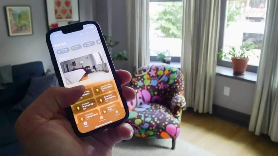 Mano che tiene un iPhone con app Casa aperta, usato per controllare luci e dispositivi smart in un salotto moderno con poltrona colorata.