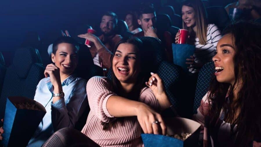 Gruppo di ragazzi e ragazze seduti al cinema mentre guardano il film, ridono e mangiano popcorn, in un clima conviviale e partecipato.