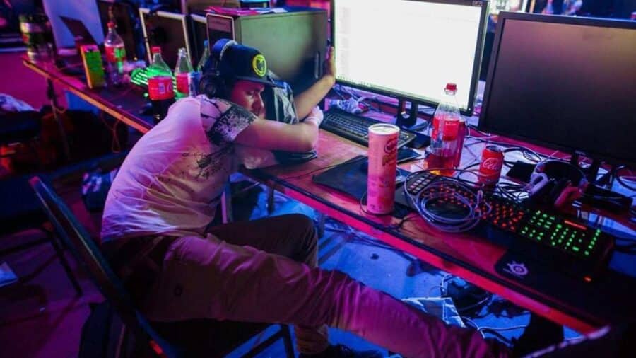 Ragazzo addormentato davanti a una postazione gaming con monitor, tastiera RGB, cuffie e bevande energetiche in una sala illuminata da luci colorate.