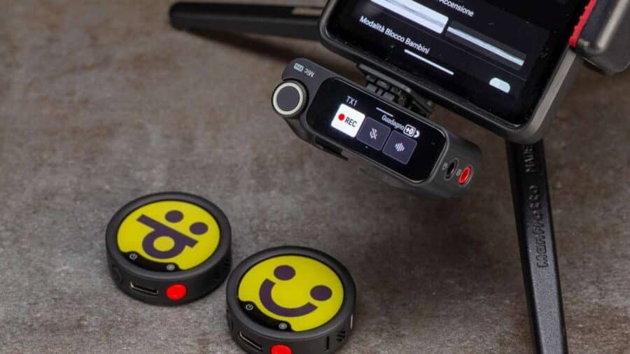 Due trasmettitori rotondi di Insta360 Mic Pro appoggiati su un tavolo accanto al ricevitore montato su una videocamera, con display acceso e interfaccia di registrazione visibile.