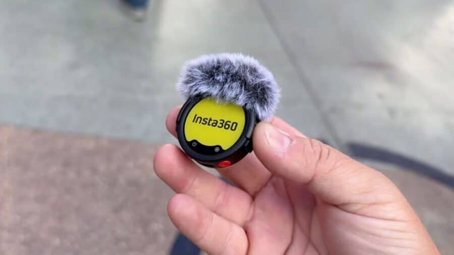 Primo piano di un trasmettitore Insta360 Mic Pro tenuto tra le dita, con filtro antivento montato sulla parte superiore e logo Insta360 visibile sul display frontale.