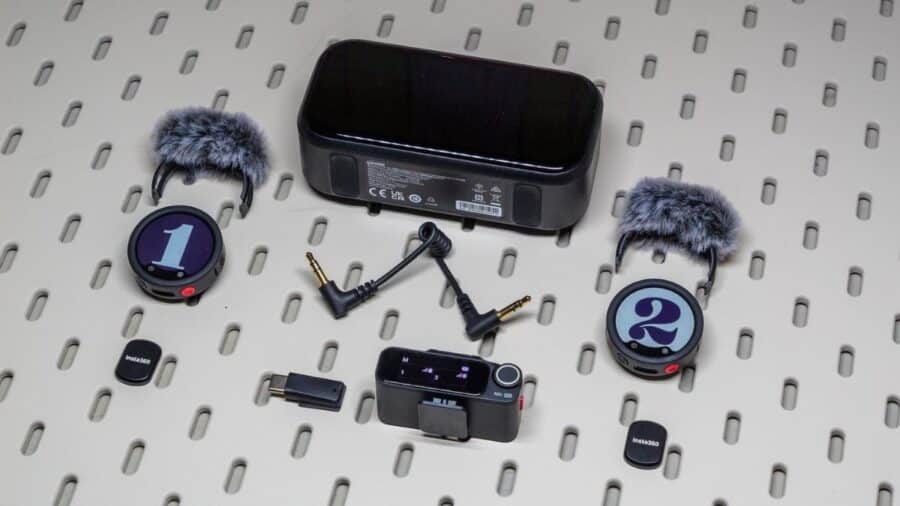 Kit completo di Insta360 Mic Pro disposto su una superficie traforata con due trasmettitori numerati, due filtri antivento, ricevitore, adattatori e custodia di ricarica.