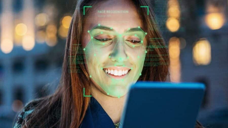 Donna sorride guardando uno smartphone mentre un’interfaccia grafica verde sovrapposta evidenzia punti del volto, simulando un sistema di riconoscimento facciale.