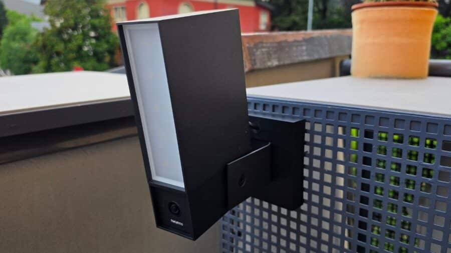 Netatmo Videocamera Esterna Original con faretto LED acceso vista frontale, installata su ringhiera in ambiente esterno luminoso