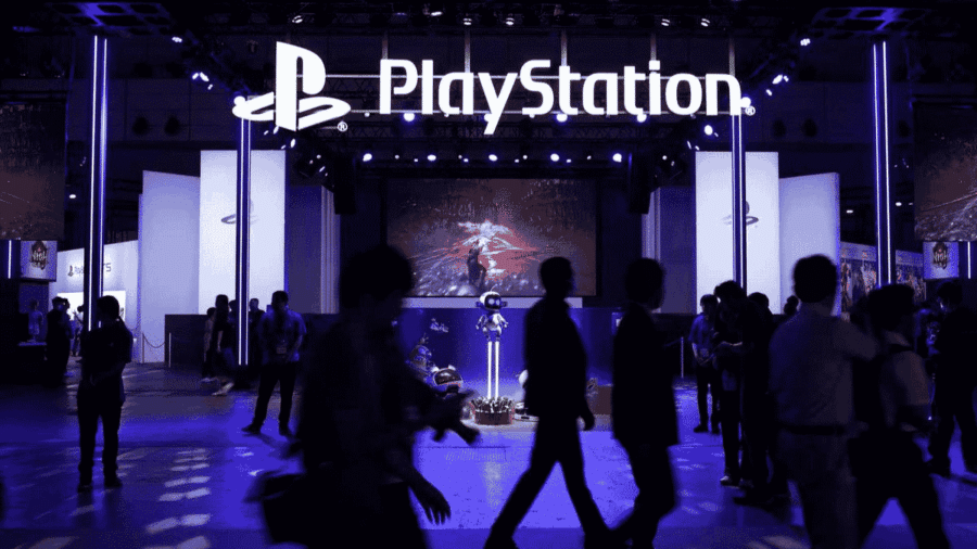 Stand PlayStation illuminato durante una fiera videoludica, con logo PlayStation in evidenza e visitatori in movimento.