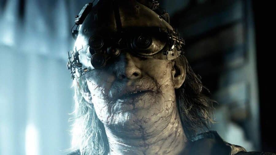 Primo piano di una creatura umanoide dal volto deformato e pallido, con una maschera metallica e lenti ottiche che coprono gli occhi in un ambiente oscuro.