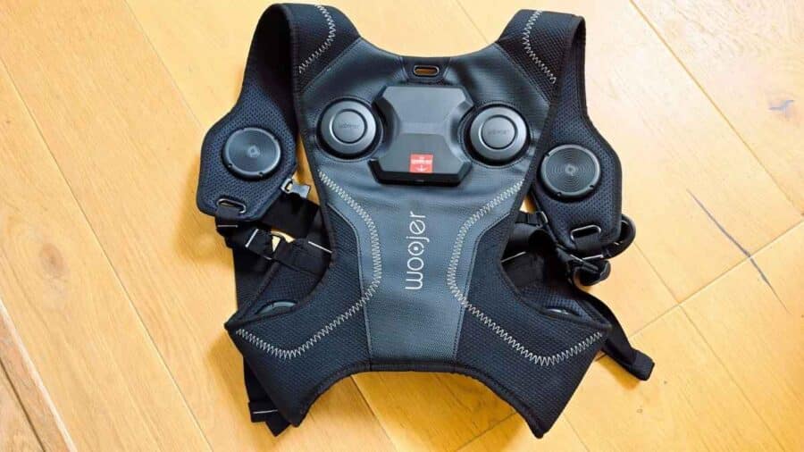 Woojer Haptic Vest 4 appoggiato su un pavimento in legno, visto dall’alto, con i trasduttori aptici e il modulo centrale ben visibili sul petto e sulle spalle.
