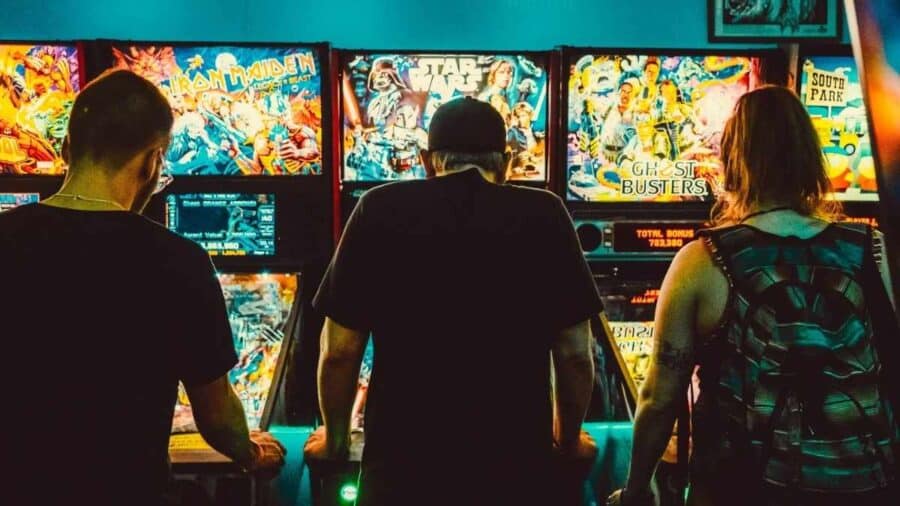 Tre persone viste di spalle giocano a flipper in una sala arcade illuminata da insegne colorate e cabine dedicate a franchise come Star Wars e Ghostbusters.