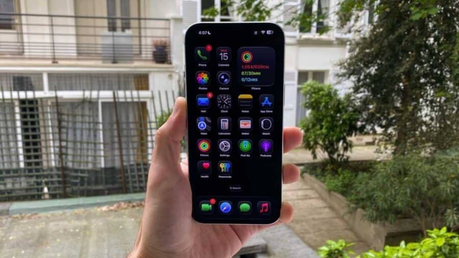 Mano che tiene un iPhone all’aperto con schermata Home in modalità scura, icone su sfondo nero e widget Fitness visibile nella parte alta del display.