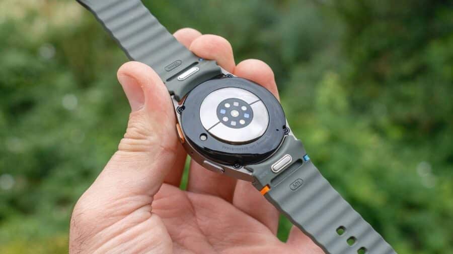 Retro di uno smartwatch sportivo con sensori biometrici a vista, tenuto in mano all’aperto, con cinturino grigio e dettagli colorati.