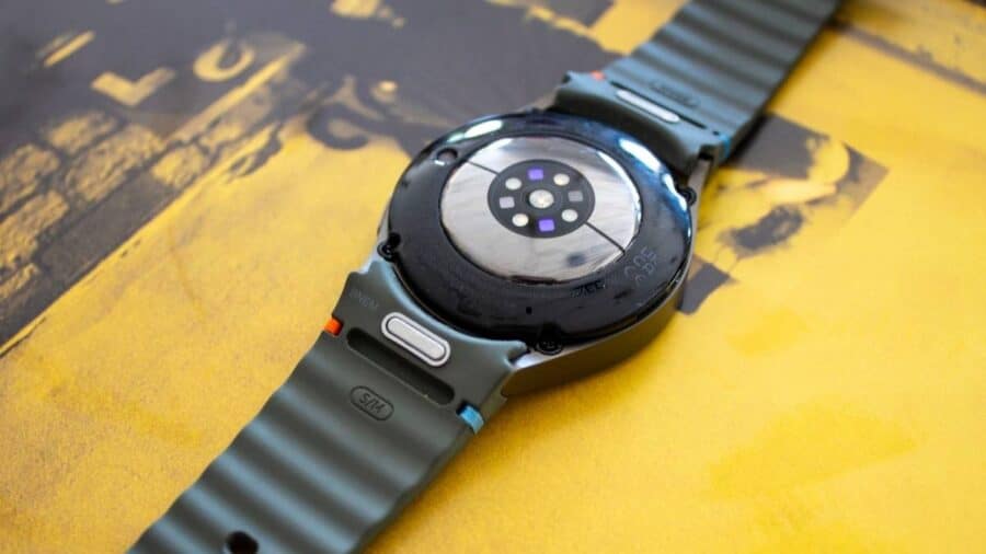 Parte posteriore di uno smartwatch appoggiato su una superficie gialla, con sensori ottici centrali e cinturino sportivo scanalato.