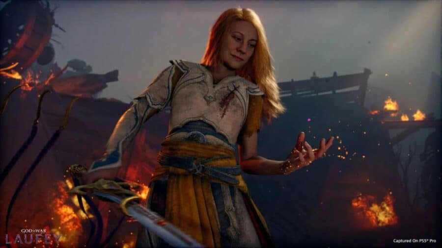Faye in God of War Laufey impugna una spada davanti a un paesaggio in fiamme, con armatura bianca e atmosfera drammatica.