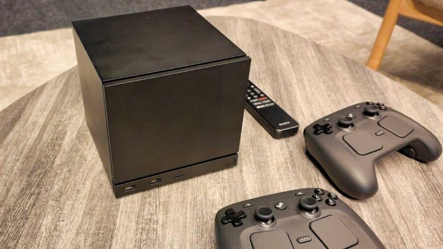 Steam Machine nera su un tavolo accanto a due controller da gioco e un telecomando Sony, in un contesto domestico da salotto.