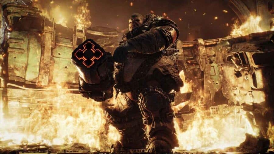 Soldato corazzato di Gears of War: E-Day avanza tra fiamme ed esplosioni con un’arma pesante in mano.
