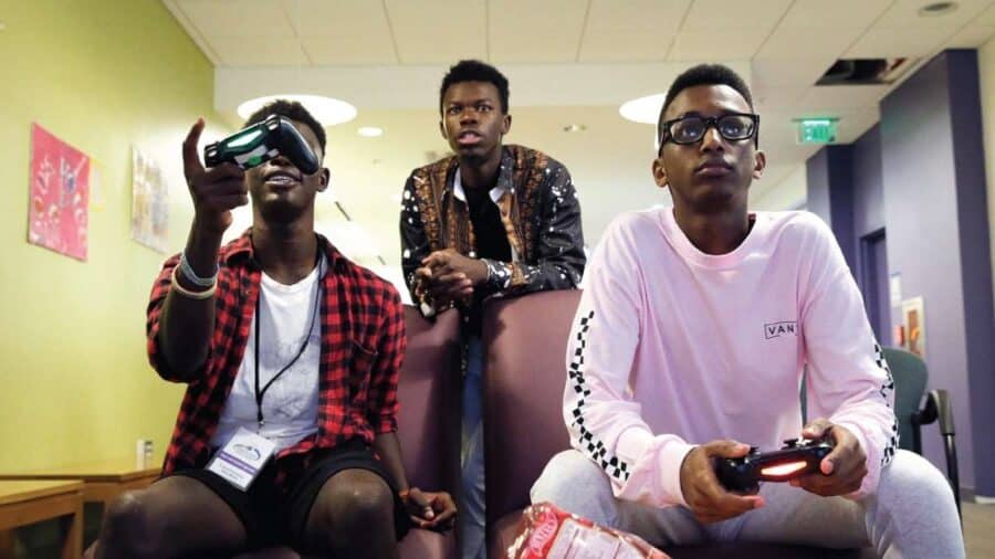 Tre ragazzi giocano ai videogiochi in una sala, con uno di loro che indossa un visore per la realtà virtuale.