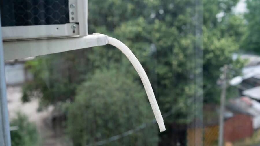 Tubo di scarico bianco di un climatizzatore che sporge verso l'esterno, dal quale fuoriesce l'acqua di condensa, con alberi verdi e vegetazione sfocata sullo sfondo.