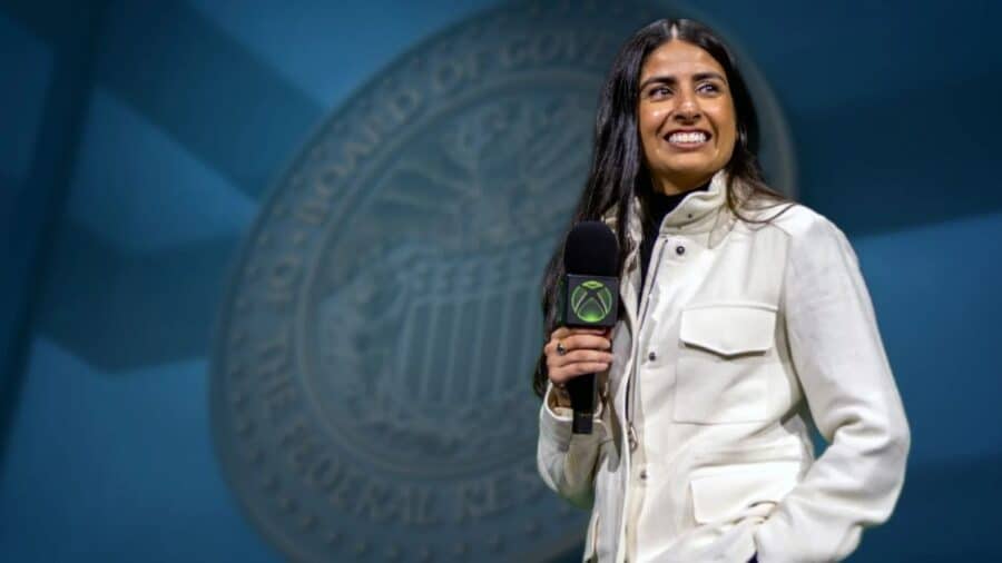 Asha Sharma sorridente impugna un microfono con il logo quadrato di Xbox; alle sue spalle campeggia l'imponente sigillo sfocato della Federal Reserve.