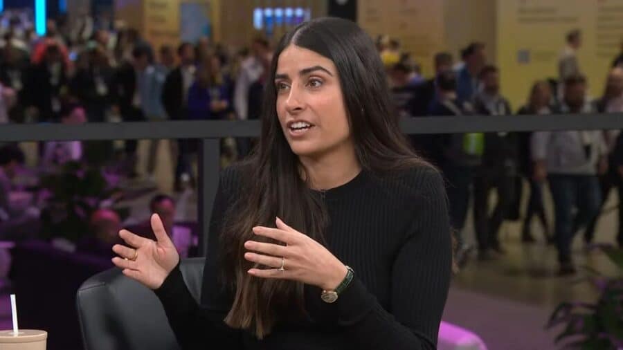 Asha Sharma, vestita con una maglia nera, parla e gesticola con le mani in primo piano mentre partecipa a un dibattito pubblico in un evento affollato.