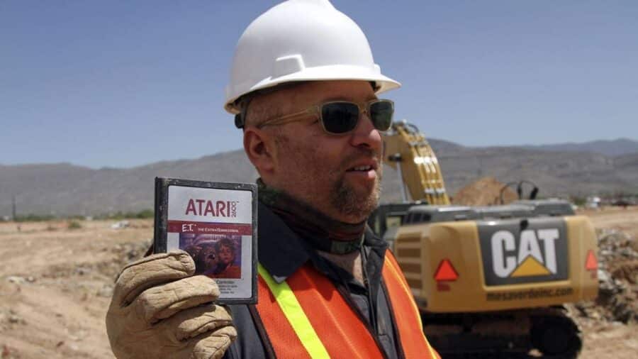 Un uomo con caschetto bianco da cantiere e occhiali da sole mostra trionfante alla telecamera una cartuccia originale di E.T. per Atari 2600 sporca di terra appena dissotterrata, con un escavatore giallo sullo sfondo.